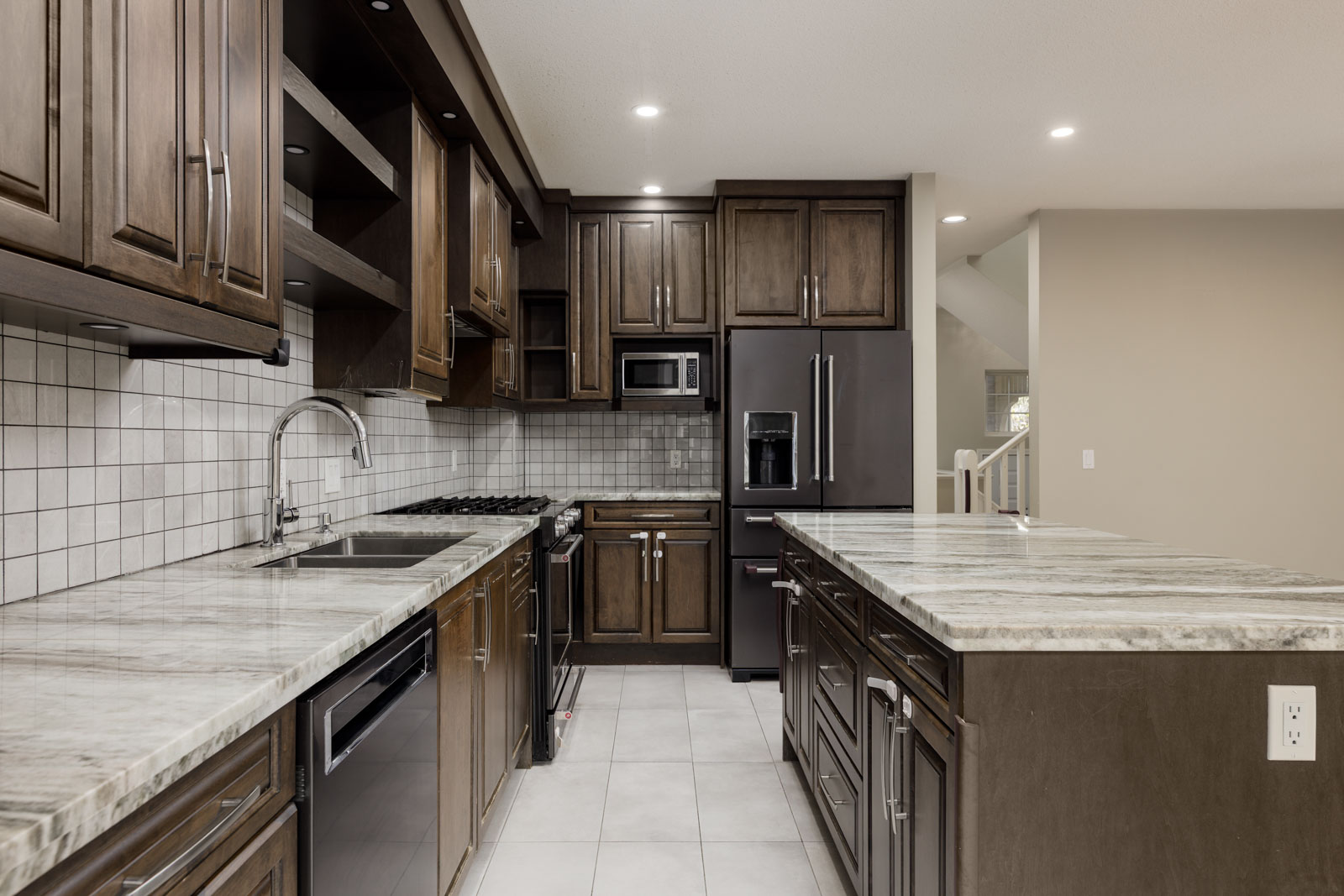 Modern kitchen with dark wood cabinets, stainless steel appliances, marble countertops, tile backsplash, and a large island under recessed lighting.