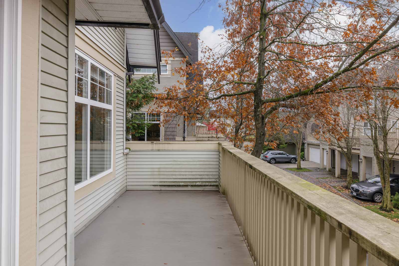 A spacious balcony with a railing overlooks a residential street with parked cars and houses; trees with autumn leaves are visible.