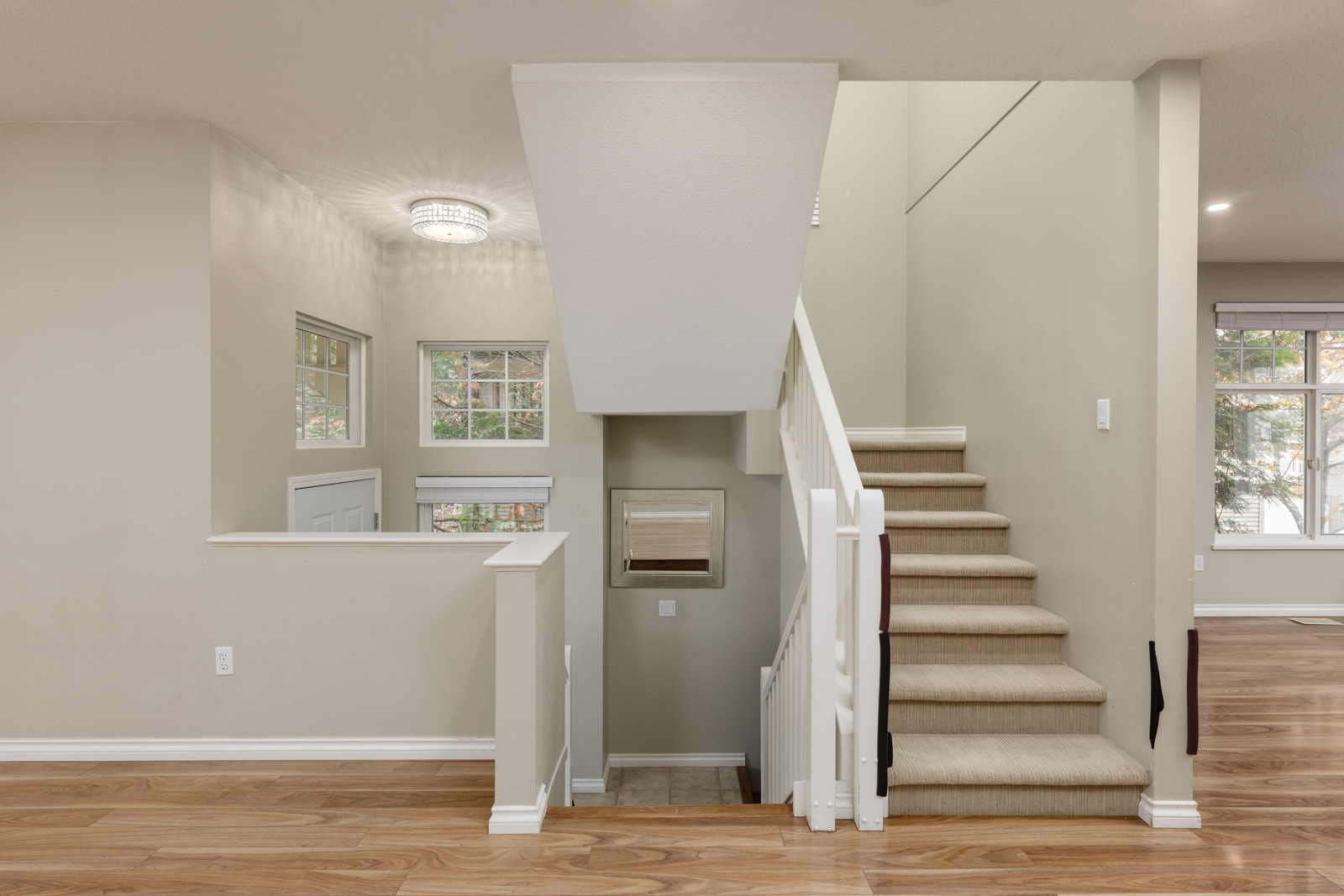 A modern interior with a carpeted staircase, white railings, light beige walls, wood flooring, and large windows letting in natural light.