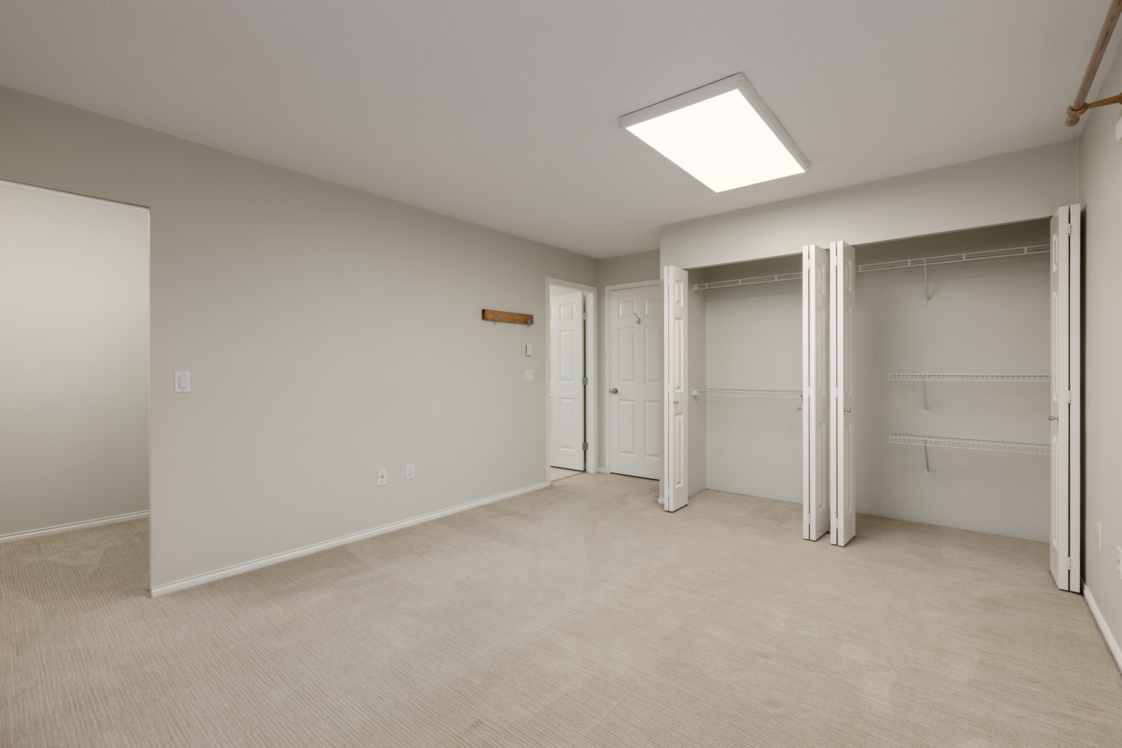 Empty bedroom with beige carpet, light gray walls, open double closet doors, wire shelving, and ceiling light panel.