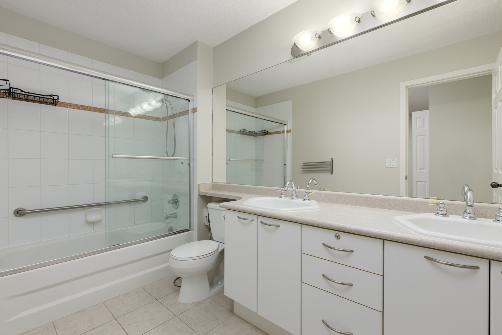Modern bathroom with a bathtub and shower, glass door, dual sinks, large mirror, white cabinets, and beige tile floor.
