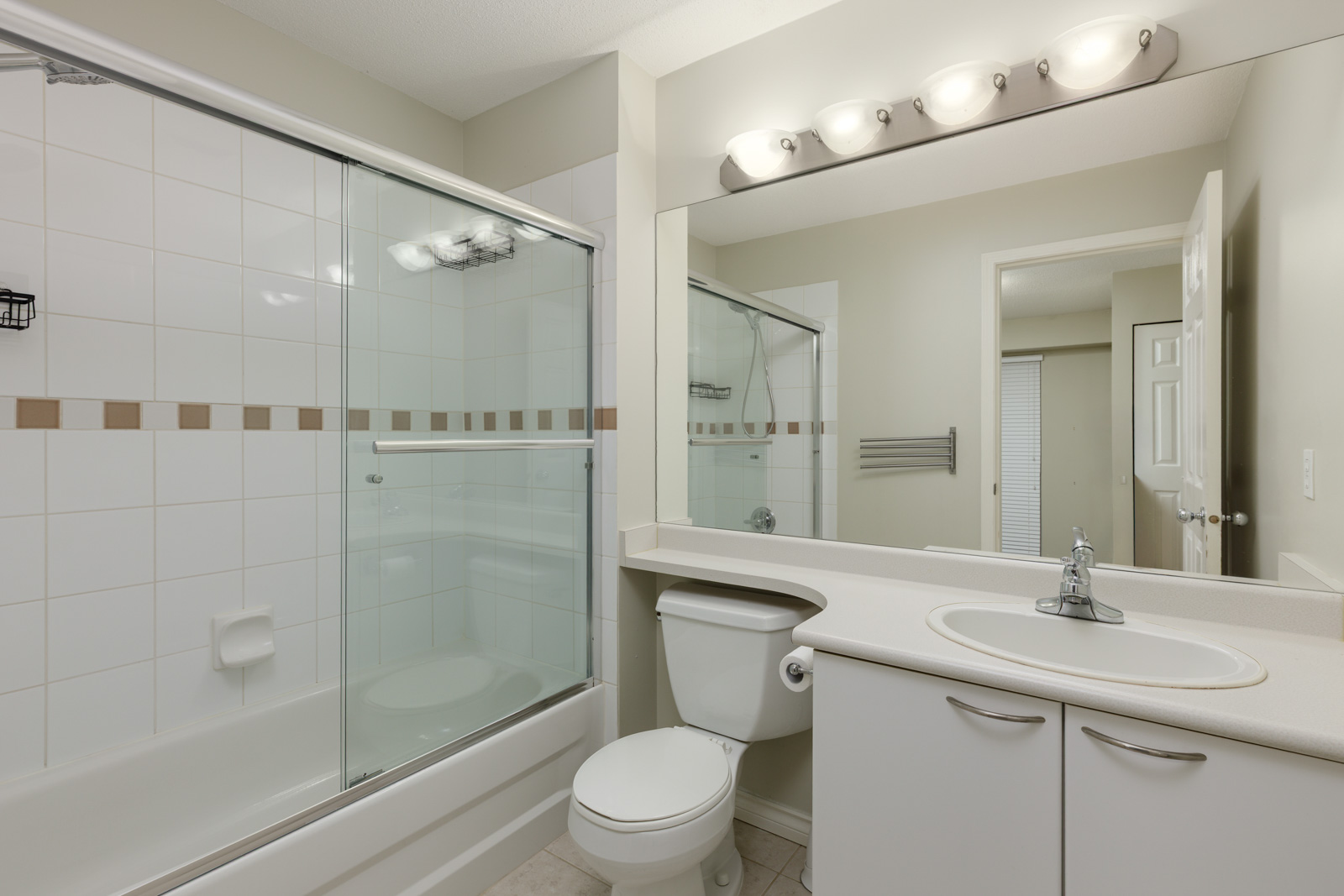 Modern bathroom with a white bathtub and glass shower doors, a toilet, a sink with cabinet, a large mirror, and wall-mounted lights above the vanity.