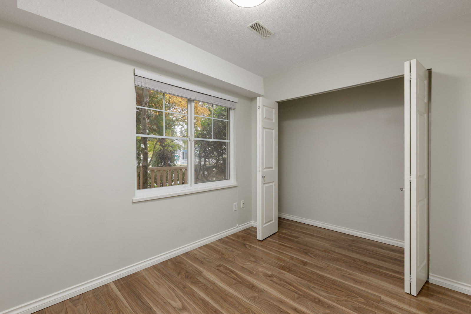 Empty room with light-colored walls, a window with a shade, open double closet doors, and wood-look flooring. Trees are visible through the window.