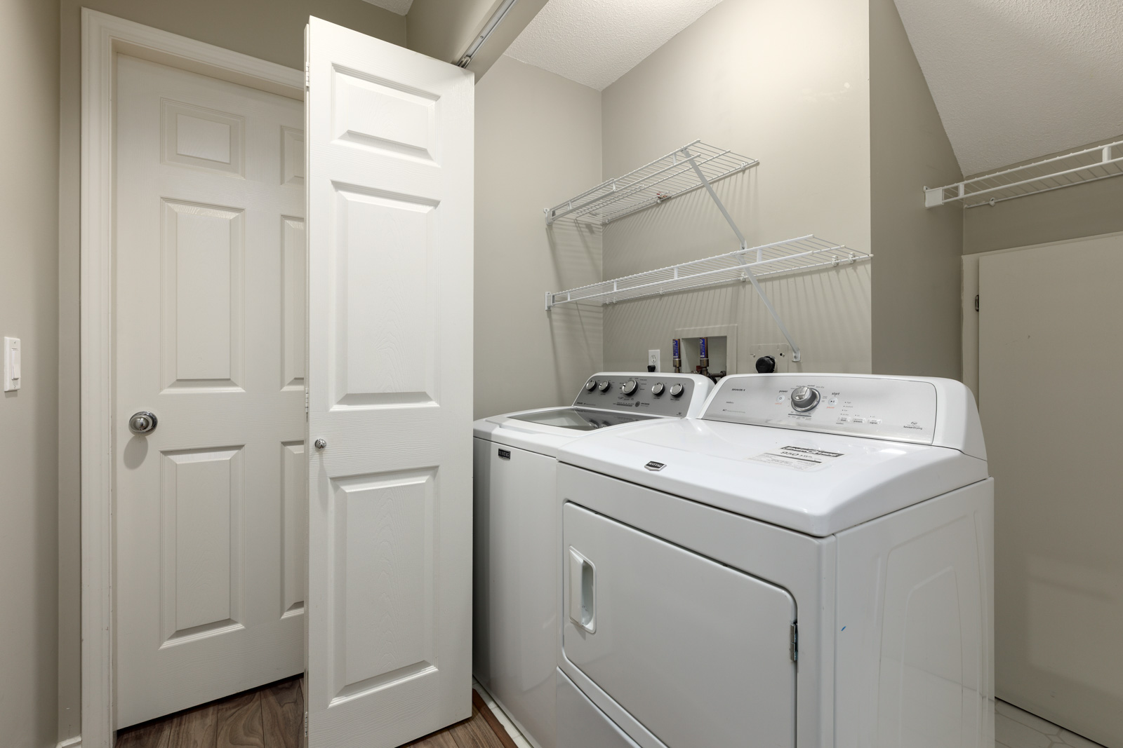 Laundry room with a washer and dryer side by side, open door, beige walls, wire shelving above the appliances, and wood-patterned flooring.