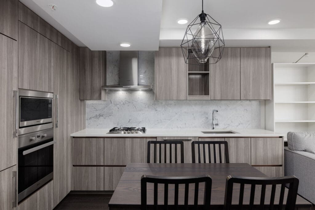 Modern kitchen with wood cabinetry, marble backsplash, built-in oven, gas cooktop, dining table with four chairs, and a geometric pendant light.