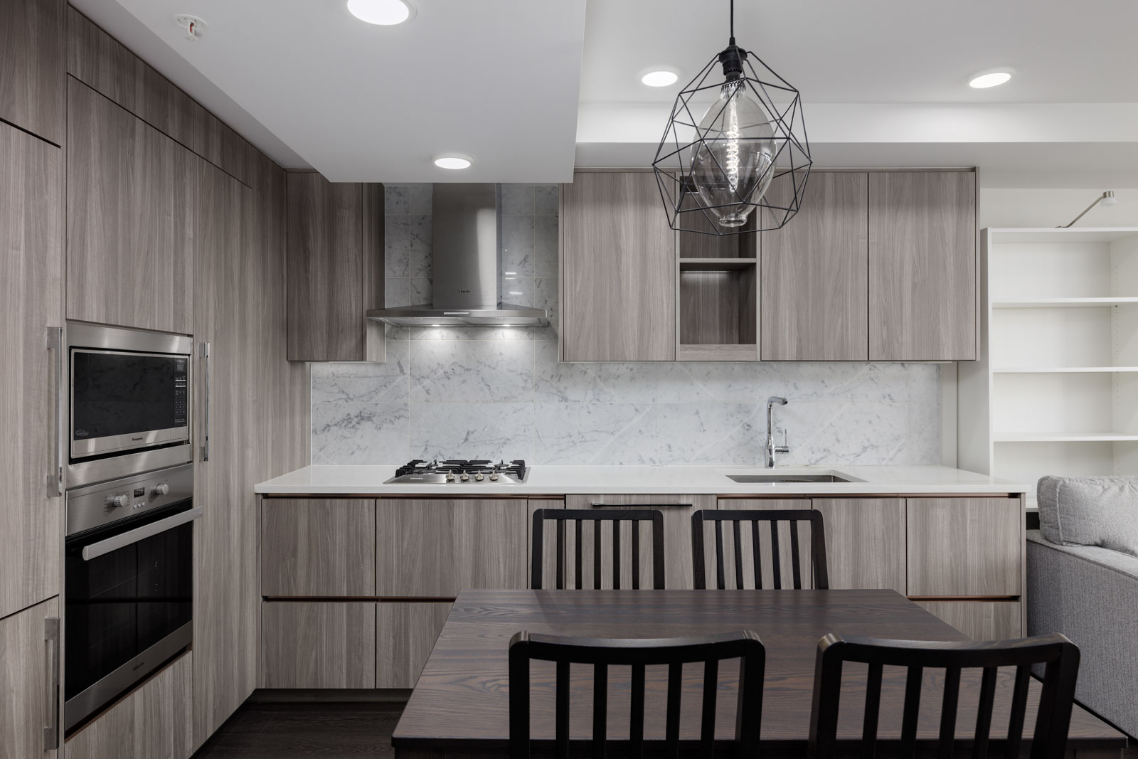 Modern kitchen with wood cabinetry, marble backsplash, built-in oven, gas cooktop, dining table with four chairs, and a geometric pendant light.