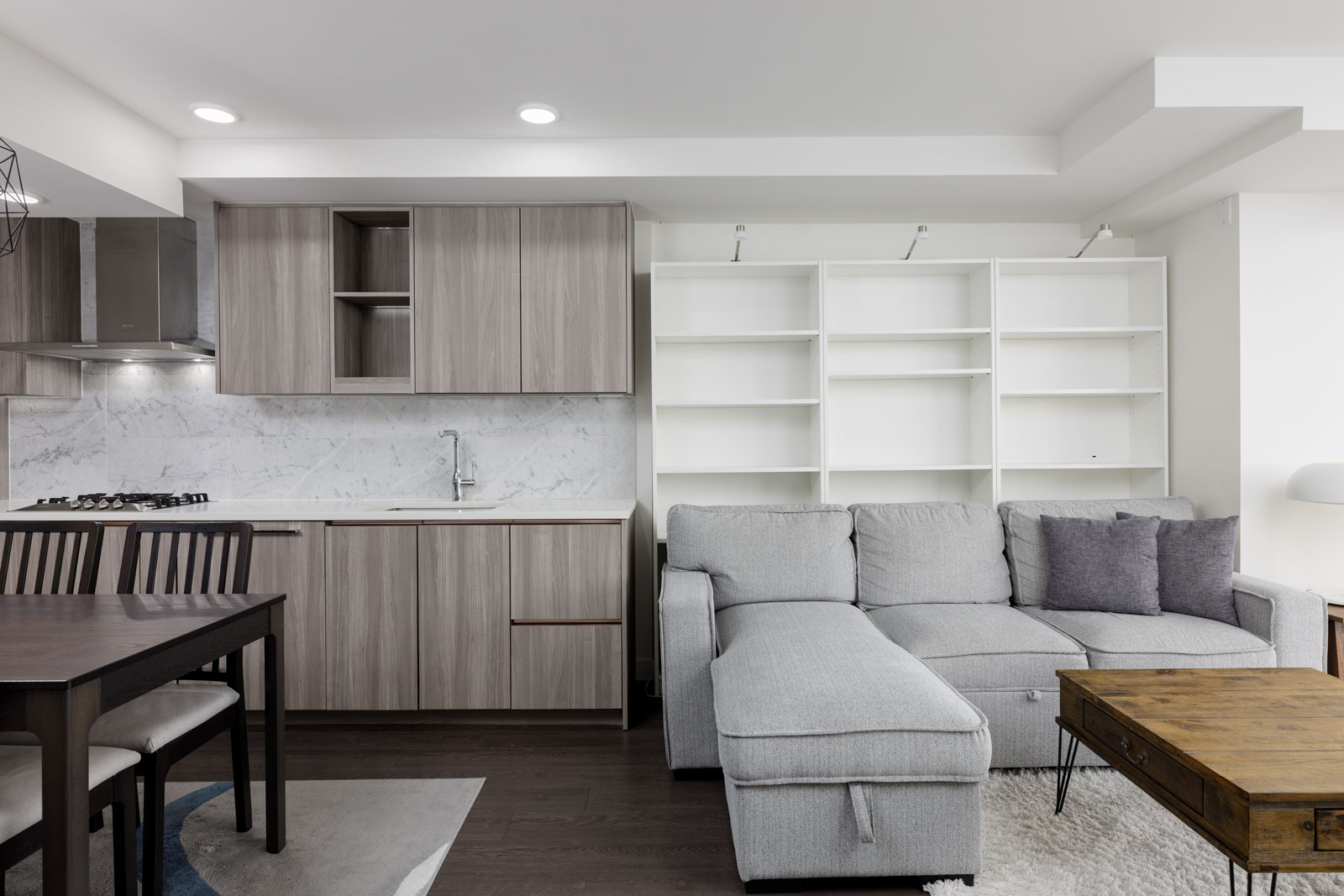 Modern open-concept living area with a gray sofa, white shelving, wooden dining table, and kitchen with marble backsplash and wood cabinets.