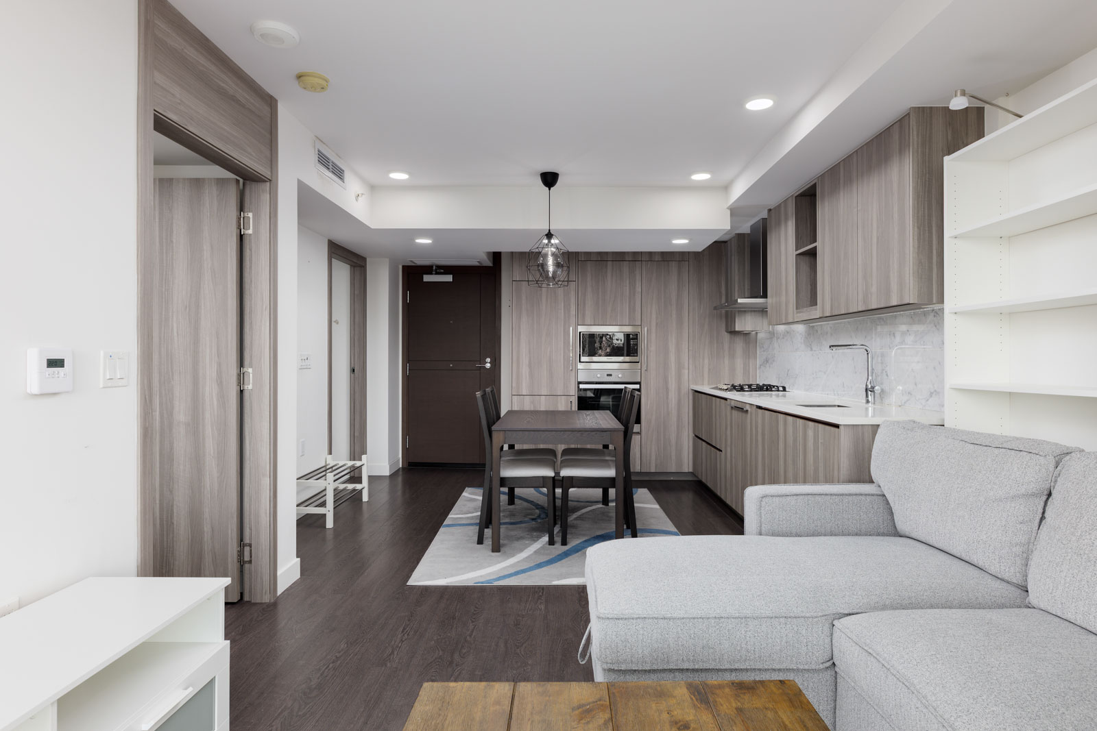 Modern apartment interior with an open-concept kitchen, dining area, light gray sectional sofa, and built-in shelves under bright lighting.