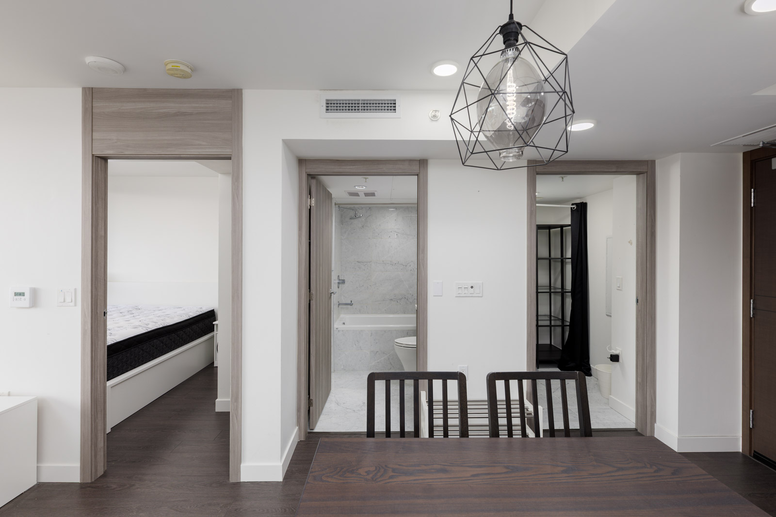 A modern apartment interior with a dining table and chairs in the foreground, and open doors leading to a bedroom, bathroom, and another room in the background.