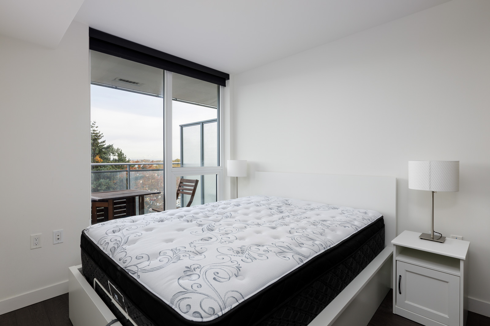 A bedroom with a large bed, white walls, a nightstand with a lamp, and a sliding glass door leading to a balcony with outdoor seating.