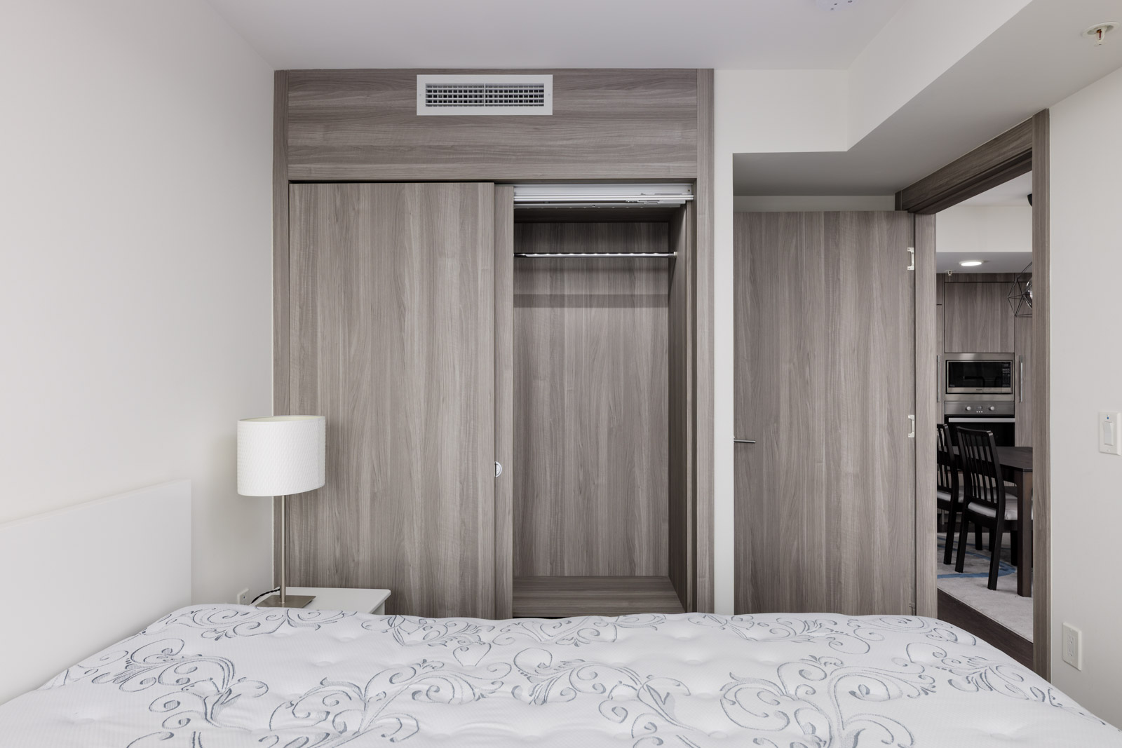 A modern bedroom with a bed in the foreground, an open closet with sliding doors, a nightstand with a lamp, and a view into a kitchen and dining area.