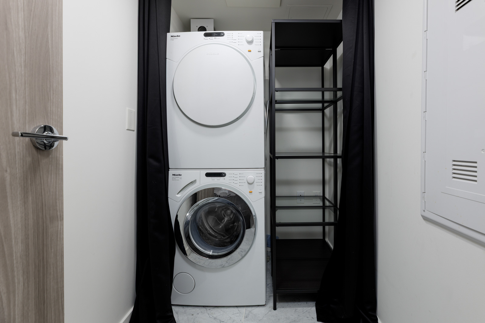 A stacked washer and dryer next to a black metal shelving unit in a small laundry closet with white walls and a wooden door.