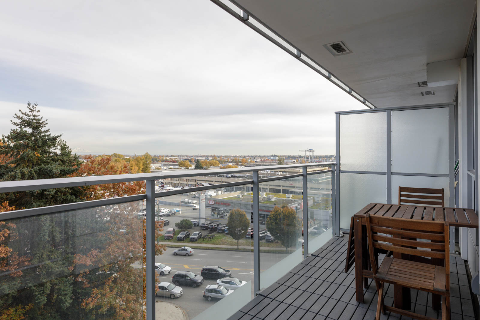 A balcony with a glass railing and wooden table and chairs overlooks a street, parking lot, and distant trees under a cloudy sky.