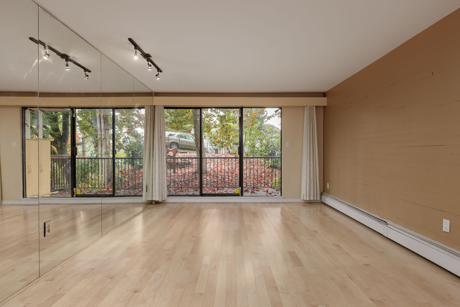 Empty room with light wood floors, mirrored wall on the left, large windows and glass doors leading to a balcony, and a brown accent wall on the right.