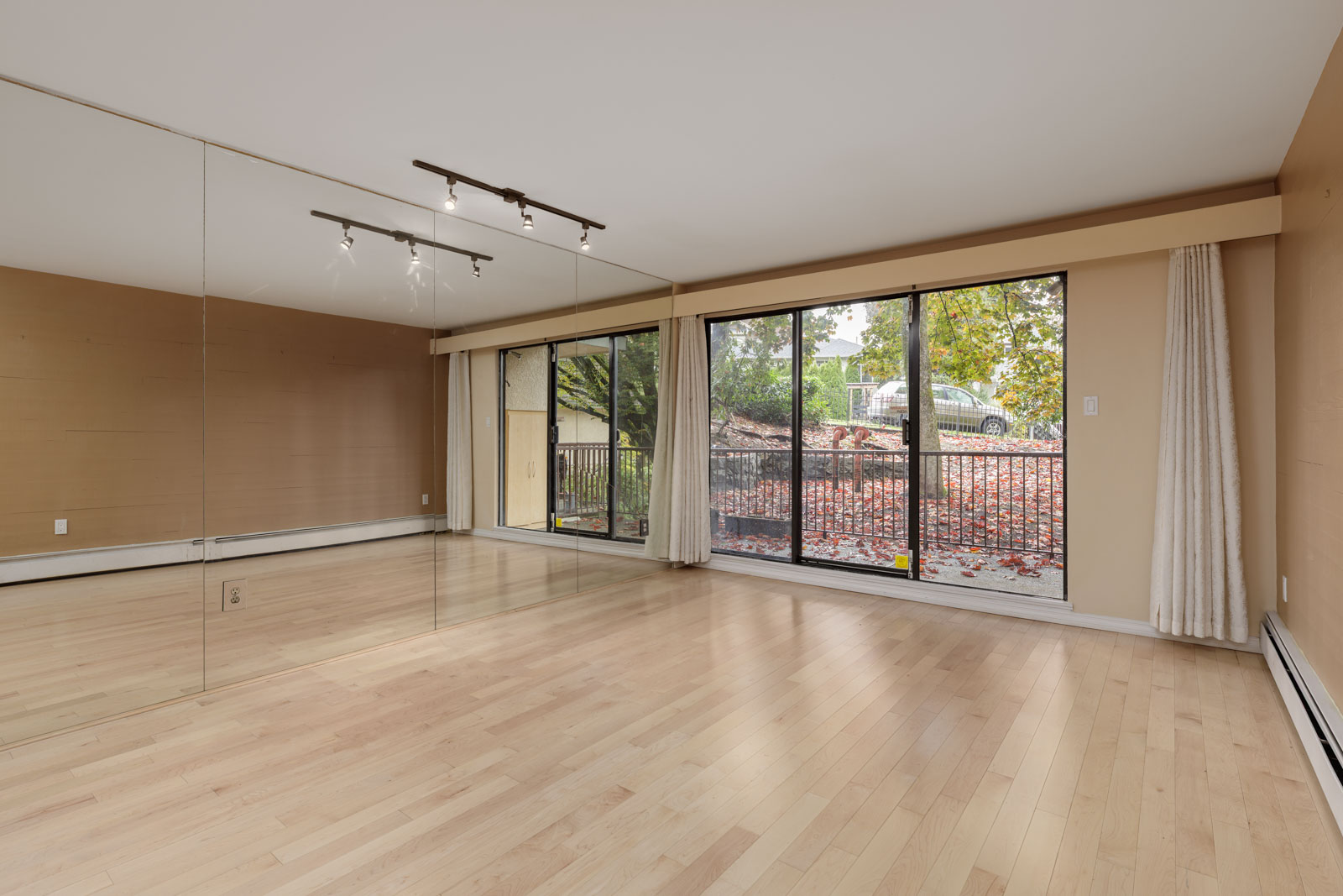 Empty room with light wood floors, mirrored wall, track lighting, and large sliding glass doors leading to a balcony with a view of trees and a parked car outside.