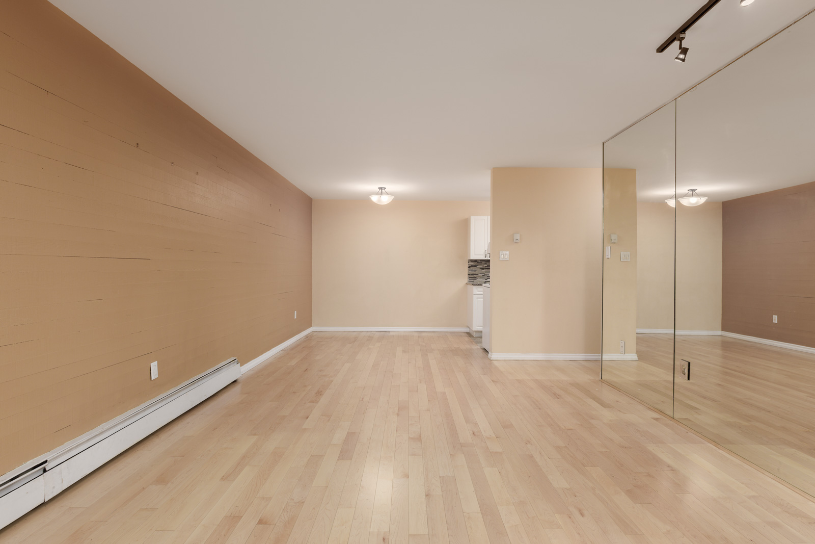Empty room with light wood flooring, tan walls, a mirrored wall on the right, and white baseboard heaters, with a partial view of a kitchen in the background.