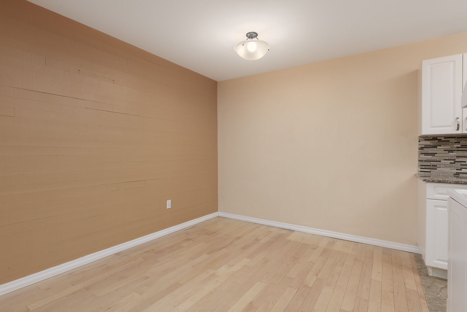 Empty room with light wood flooring, beige and tan walls, a ceiling light fixture, and part of a kitchen countertop and cabinets visible on the right.