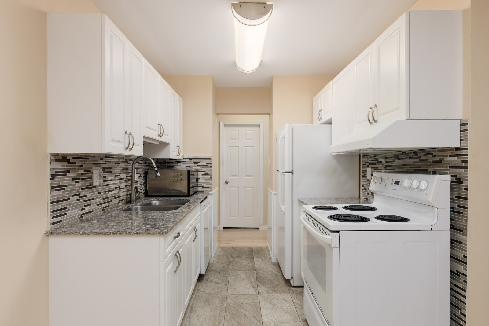 A small kitchen with white cabinets, granite countertops, a tile backsplash, white appliances, and a tiled floor, viewed facing a closed door.