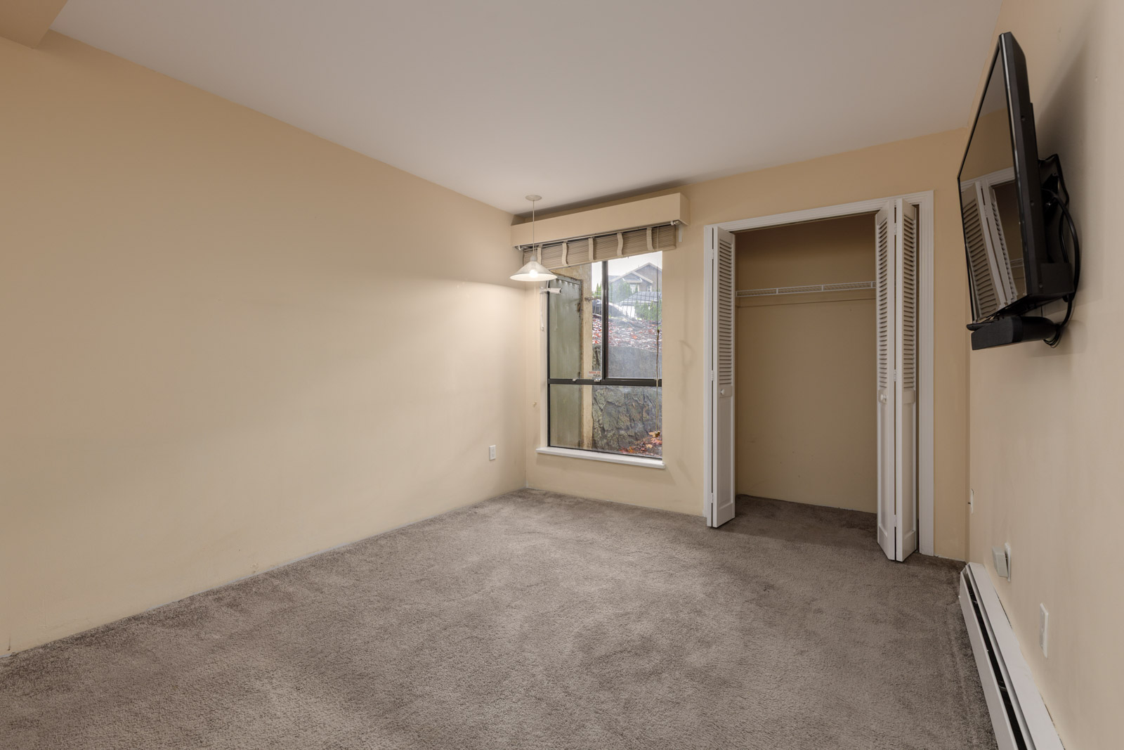 Empty beige-walled bedroom with carpeted floor, open closet, wall-mounted TV, and a window with a view outside. Ceiling light fixture hangs near the window.