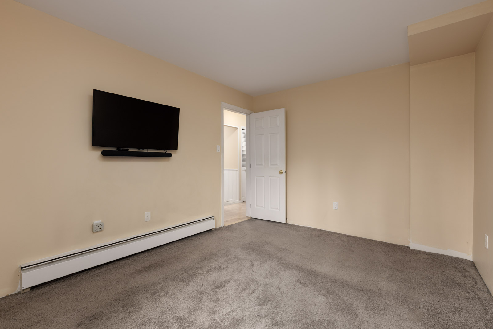 Empty room with beige walls, gray carpet, a wall-mounted TV, baseboard heater, and an open door leading to a lit hallway.