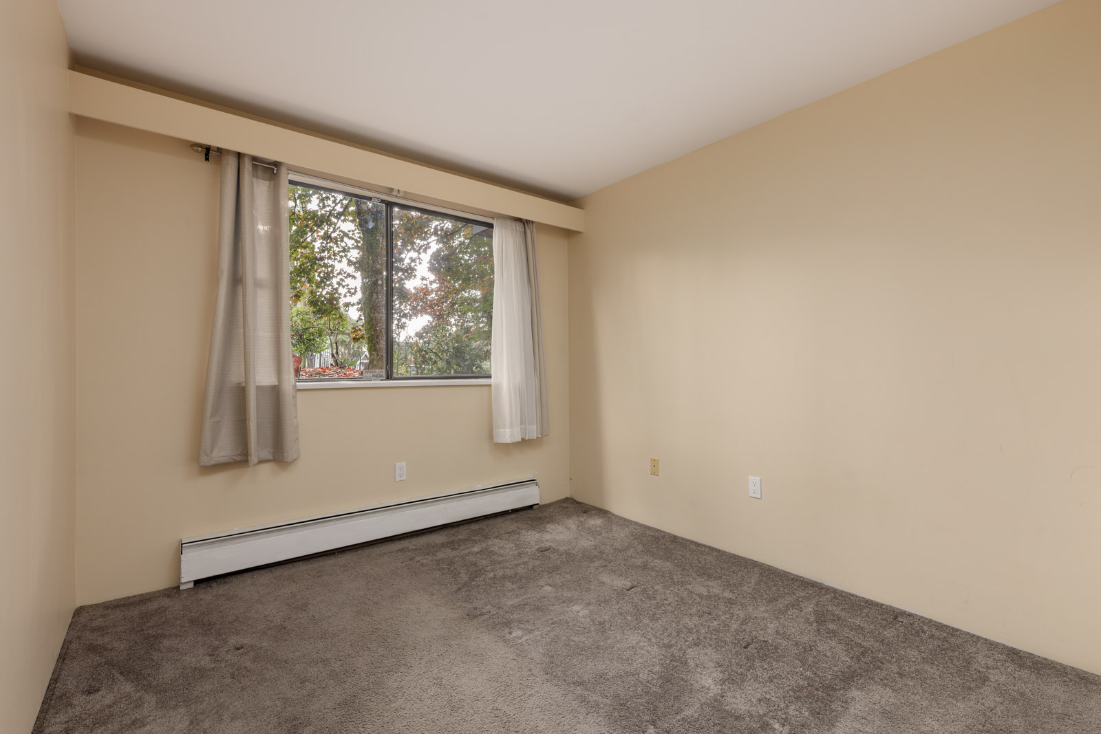 Empty room with beige walls, grey carpet, a window with sheer curtains, and a white baseboard heater below the window.