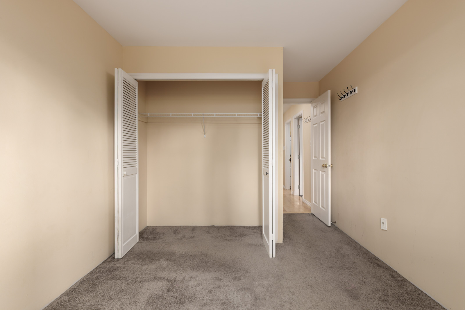 Empty beige room with open closet doors, gray carpet flooring, a white door leading to a hallway, and wall-mounted coat hooks.