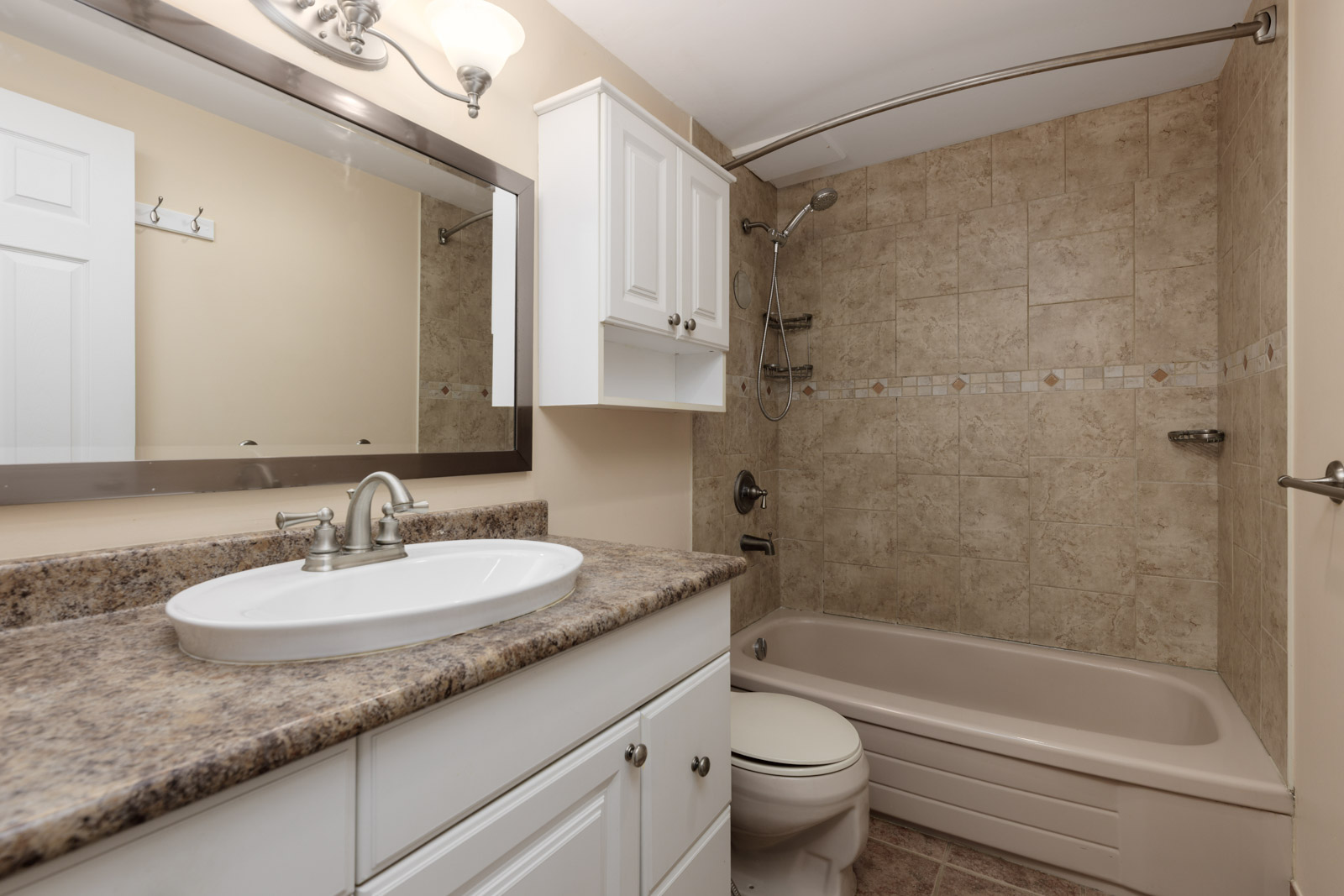 A bathroom with a granite countertop, white vanity, sink, mirror, toilet, and a beige-tiled bathtub with a showerhead and overhead light.