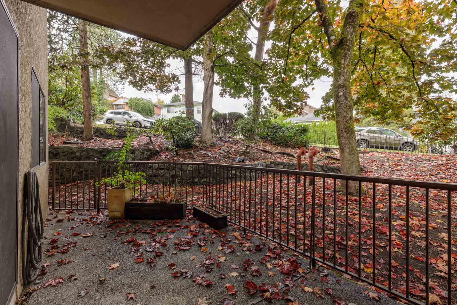 View from a covered patio with fallen autumn leaves on the ground, overlooking a sloped yard, trees, and parked cars on the street beyond a metal railing.