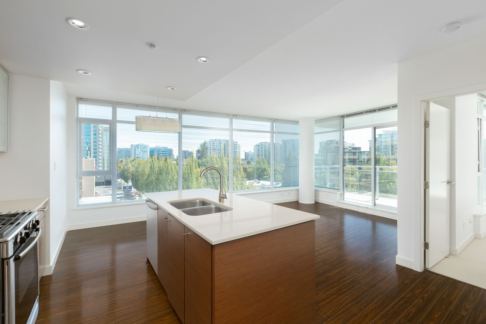 Modern, unfurnished apartment kitchen and living area with large windows, city views, and wood flooring; an island with a sink is centered in the space.