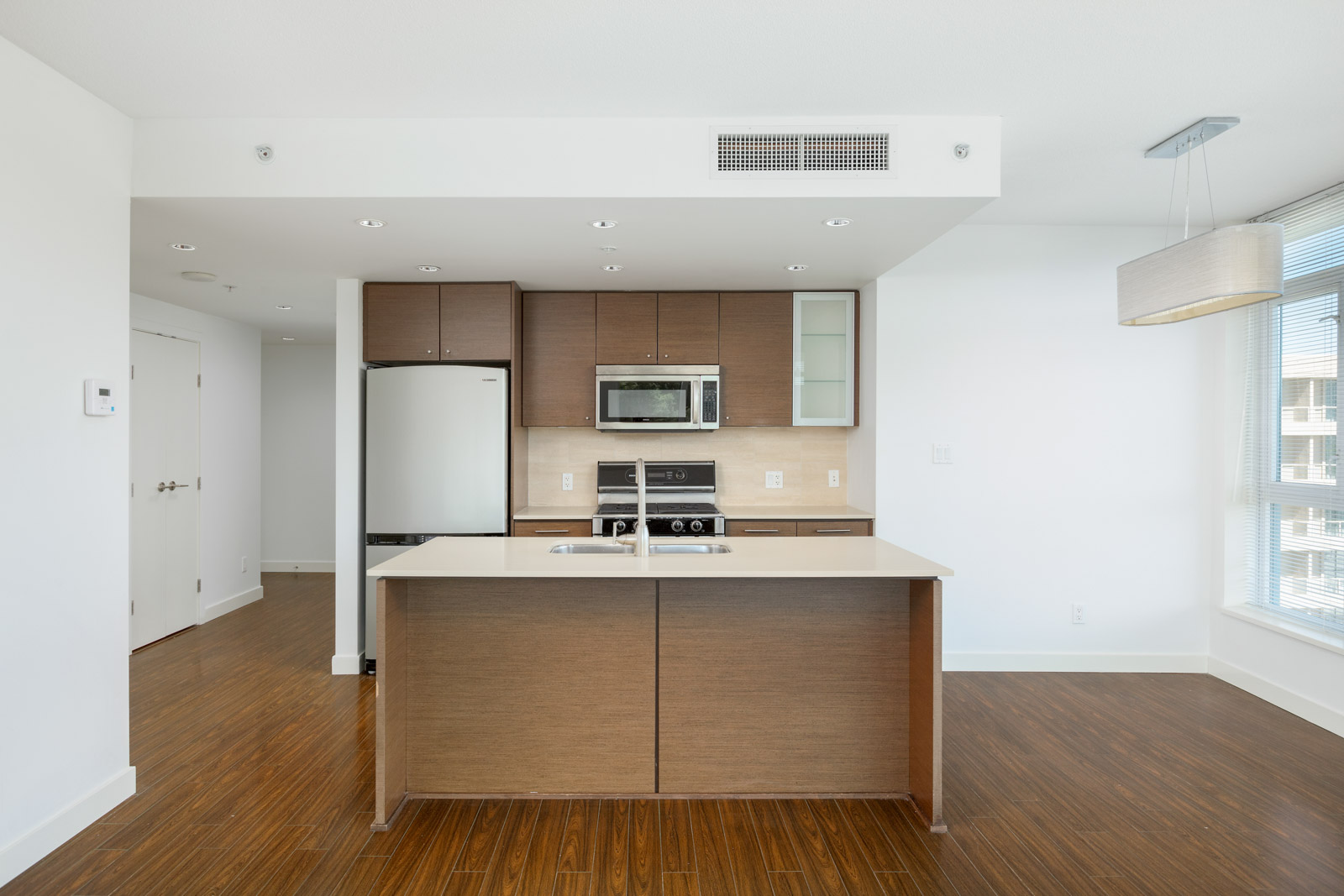 Modern kitchen with brown cabinets, stainless steel appliances, island with sink, and wood flooring in a bright, open-plan apartment.