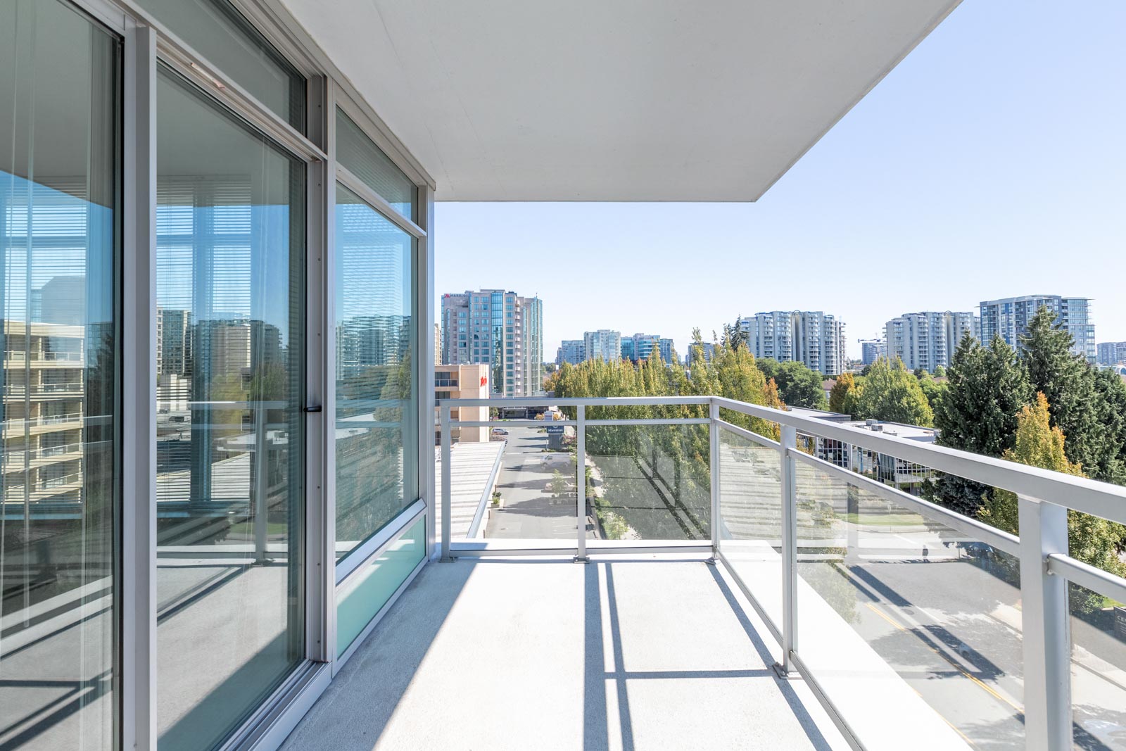 A modern apartment balcony with glass railings overlooks a cityscape with multiple high-rise buildings and trees under a clear sky.