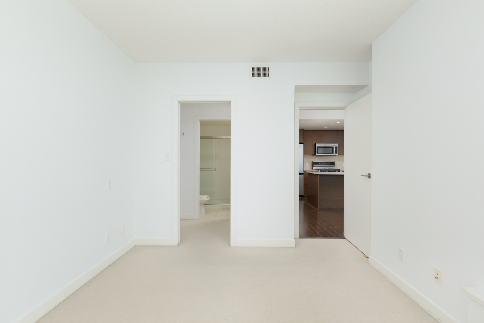 A clean, empty white room with beige carpet, two open doorways—one leading to a bathroom and one to a modern kitchen area.