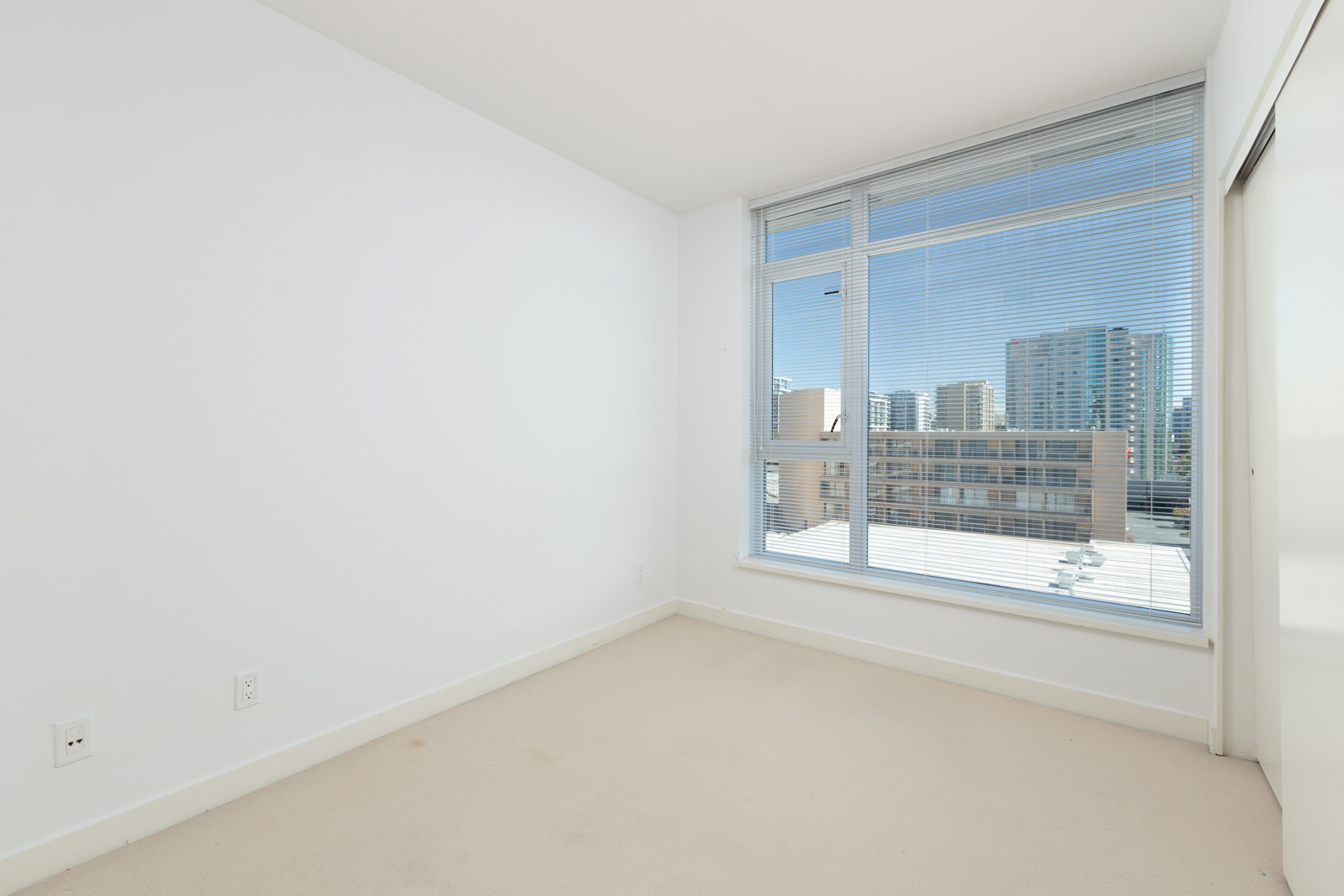 Empty room with white walls, beige carpet, and a large window with blinds overlooking city buildings under a clear sky.