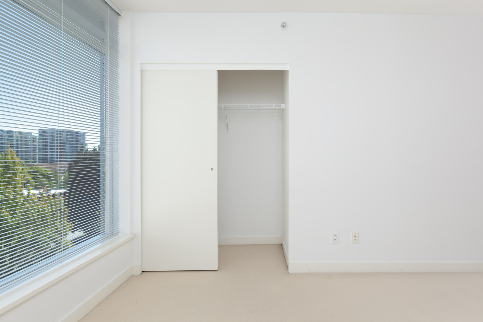 Empty white closet with sliding door in a bright room, large window with blinds on the left, overlooking trees and buildings.