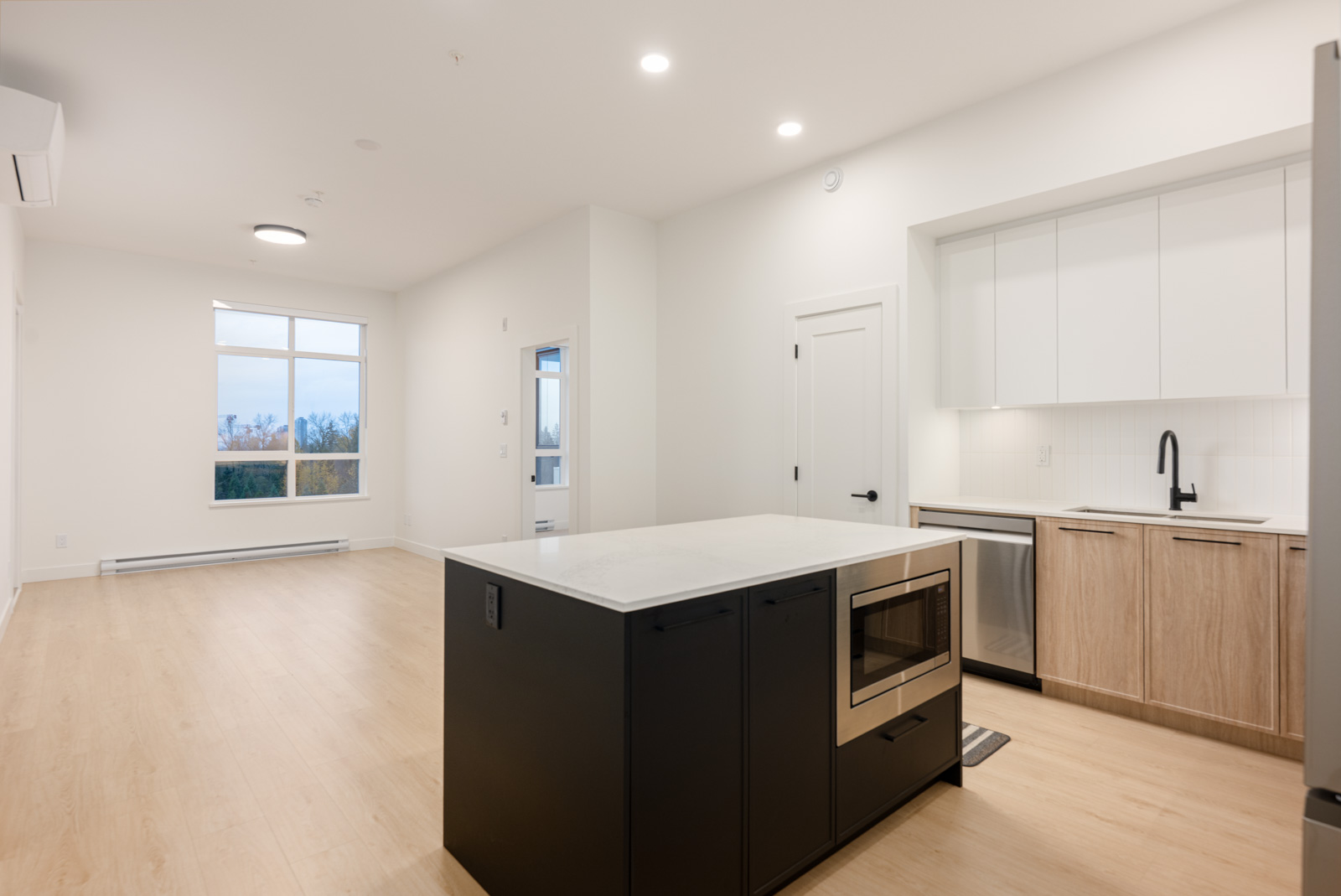 Modern open-concept kitchen and living area with light wood floors, white walls, island with built-in microwave, and large windows.
