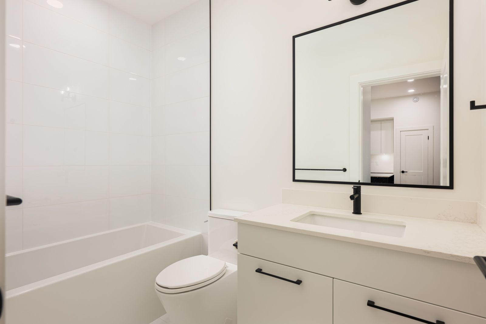 Modern bathroom with white walls, bathtub, toilet, single-sink vanity with black fixtures, and a large rectangular mirror reflecting a door.