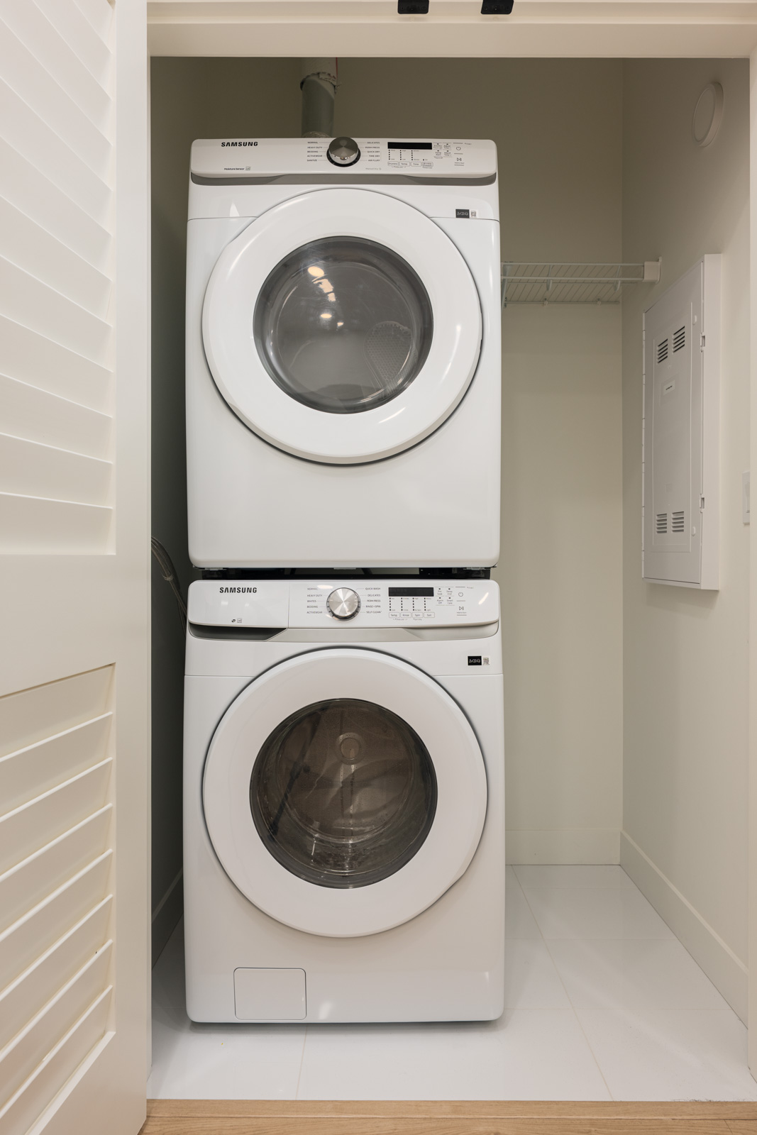 A stacked white washer and dryer unit is placed in a small laundry closet with a shelf above and an electrical panel on the right wall.