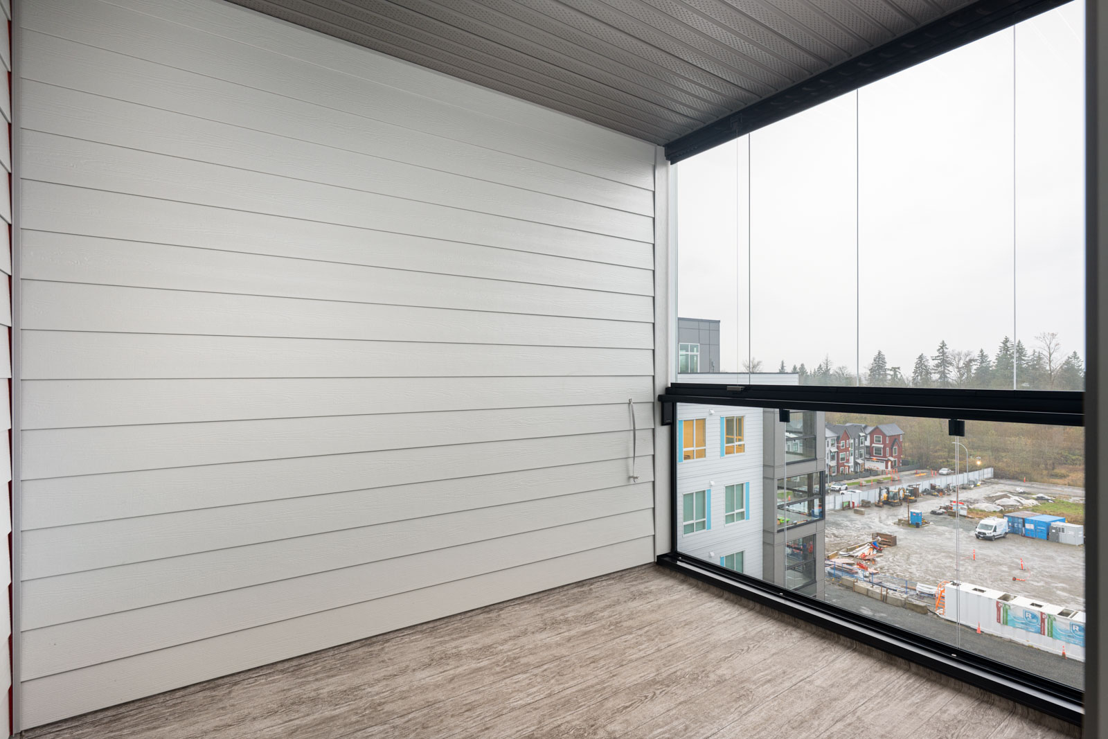 A covered balcony with a glass railing overlooks a construction site and nearby buildings on an overcast day.
