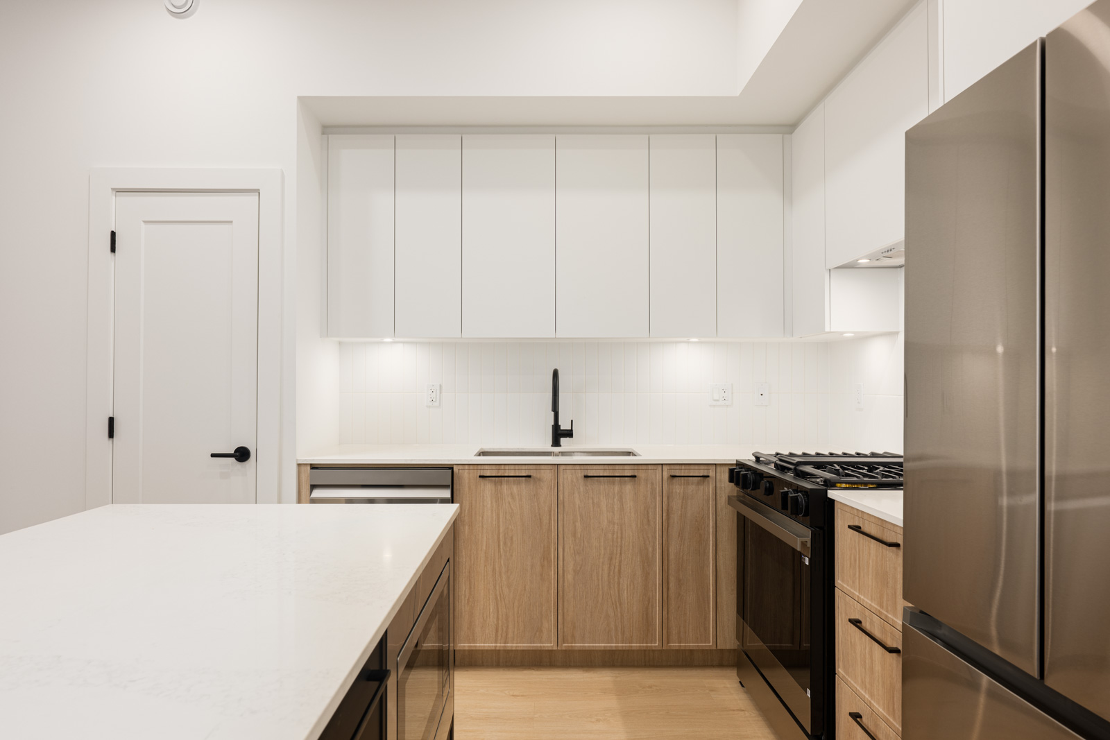 Modern kitchen with white upper cabinets, light wood lower cabinets, a stainless steel refrigerator and oven, white countertops, and a central island.