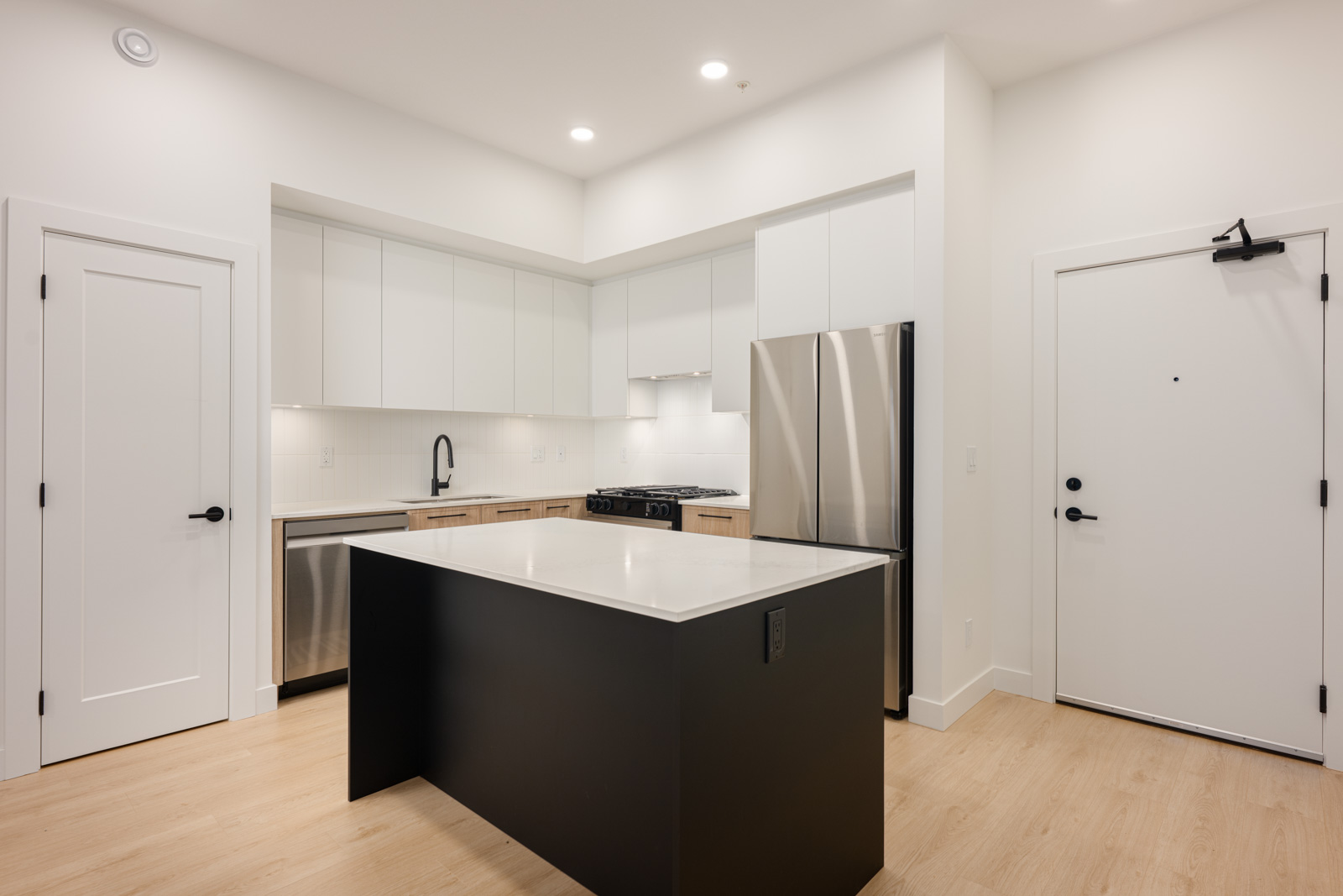 Modern kitchen with white cabinets, stainless steel appliances, a black island with a white countertop, light wood flooring, and white walls.