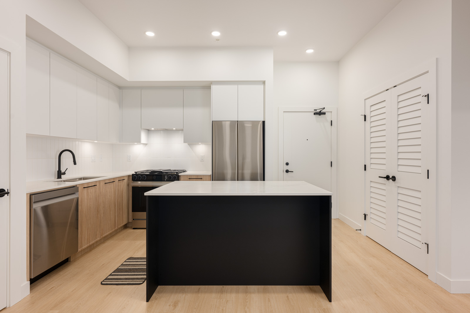 Modern kitchen with light wood floors, white cabinets, stainless steel appliances, black island, and recessed lighting. A closed door and louvered closet doors are visible in the background.