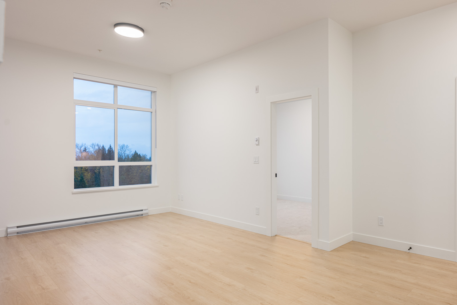 Unfurnished room with light wood flooring, white walls, a large window, baseboard heater, and an open doorway leading to another room.