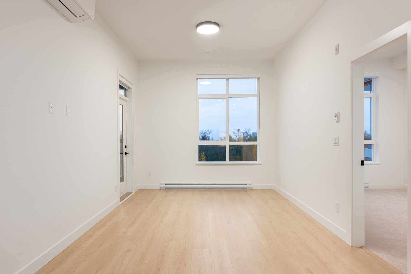 Unfurnished, modern room with light wood flooring, white walls, large window, and door to a balcony; adjacent room visible through open doorway.