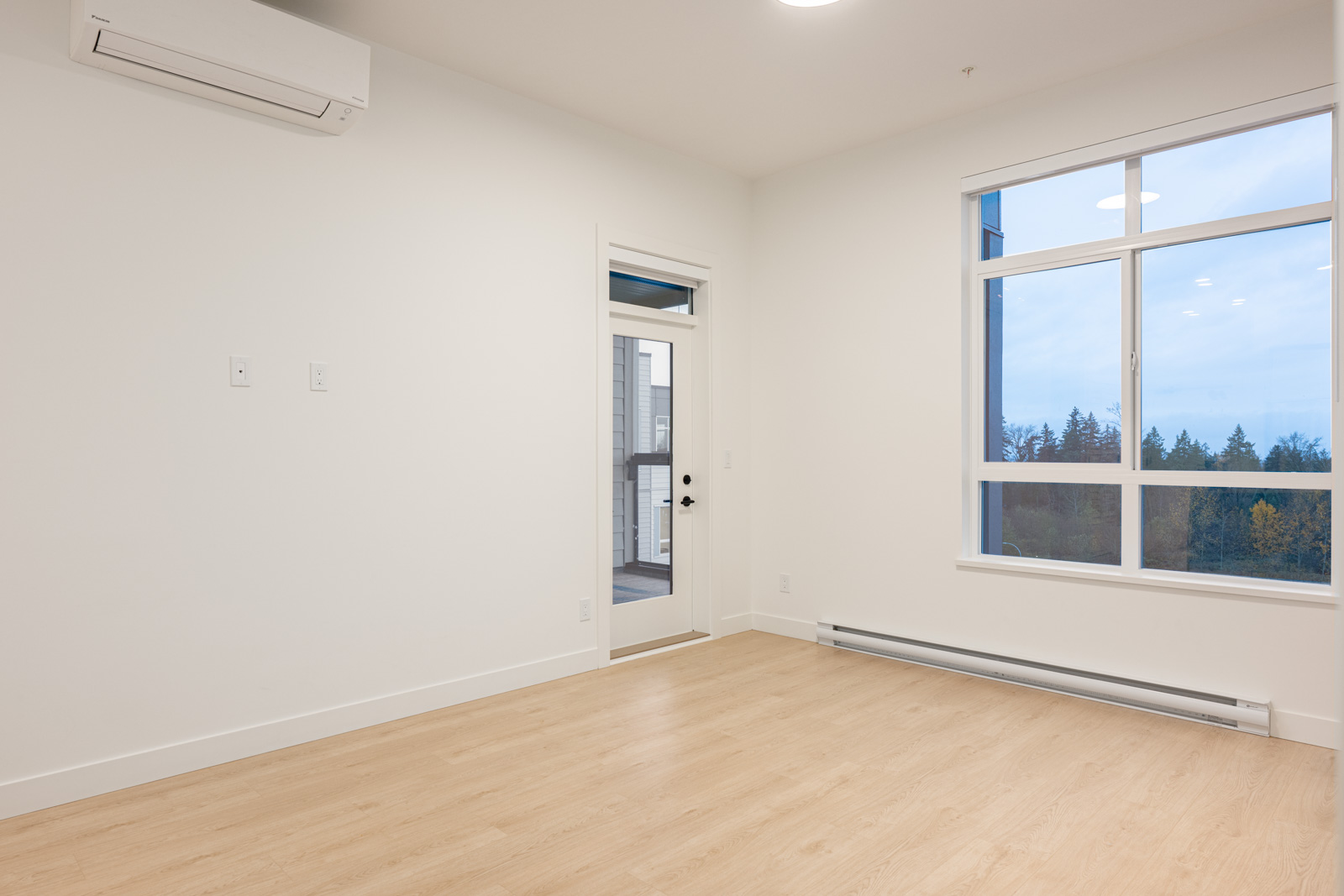 Empty room with light wood flooring, white walls, a large window, a glass door leading to a balcony, an air conditioning unit, and a baseboard heater.