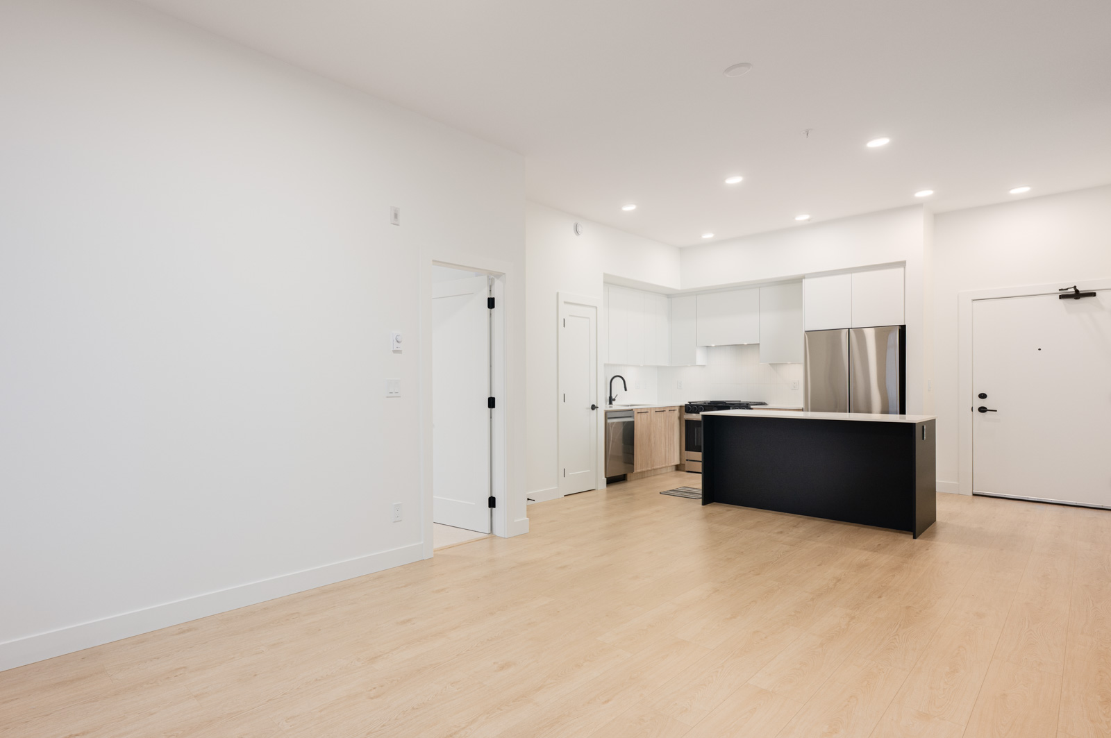 A modern, unfurnished apartment interior with light wood floors, white walls, and a kitchen featuring white cabinets, stainless steel appliances, and a black island.