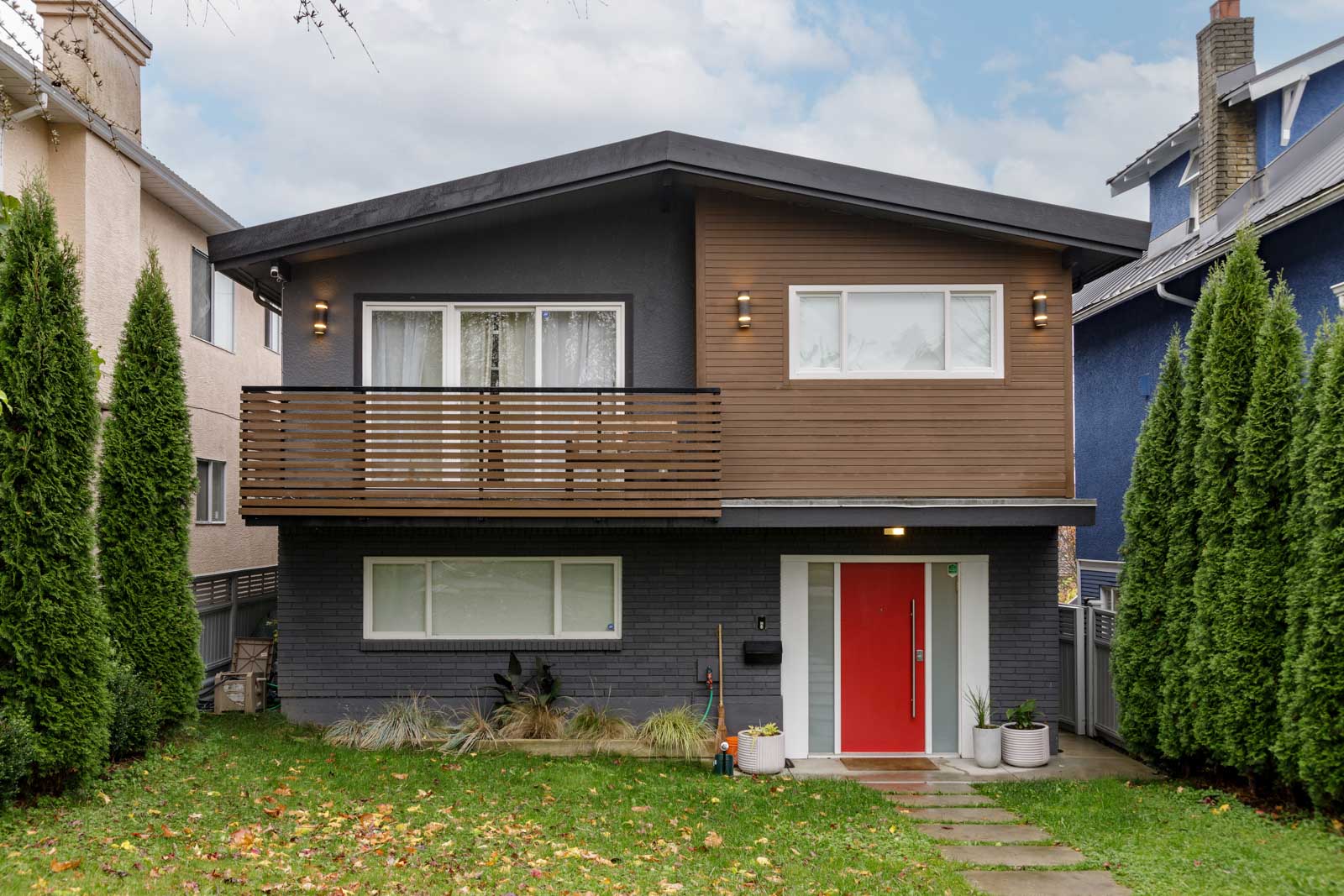 Two-story modern house with dark gray and brown exterior, a red front door, balcony with wooden slats, and surrounded by tall evergreen trees on both sides.
