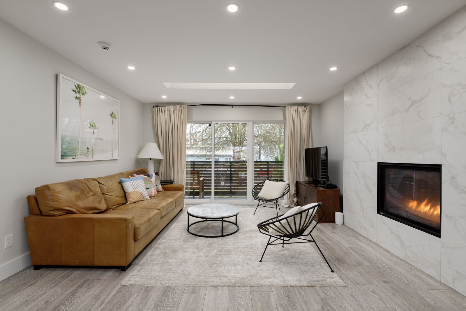Modern living room with a tan sofa, round coffee table, accent chairs, wall-mounted TV, fireplace, large artwork, and sliding glass doors leading to a balcony.