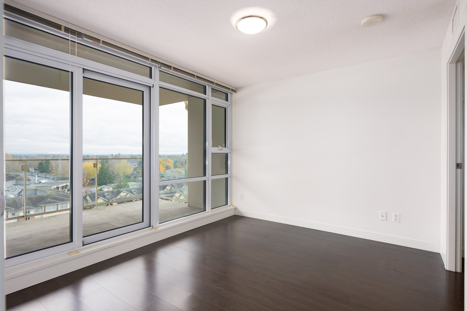 Empty room with large floor-to-ceiling windows, dark wood floors, white walls, and a view of buildings and trees outside.