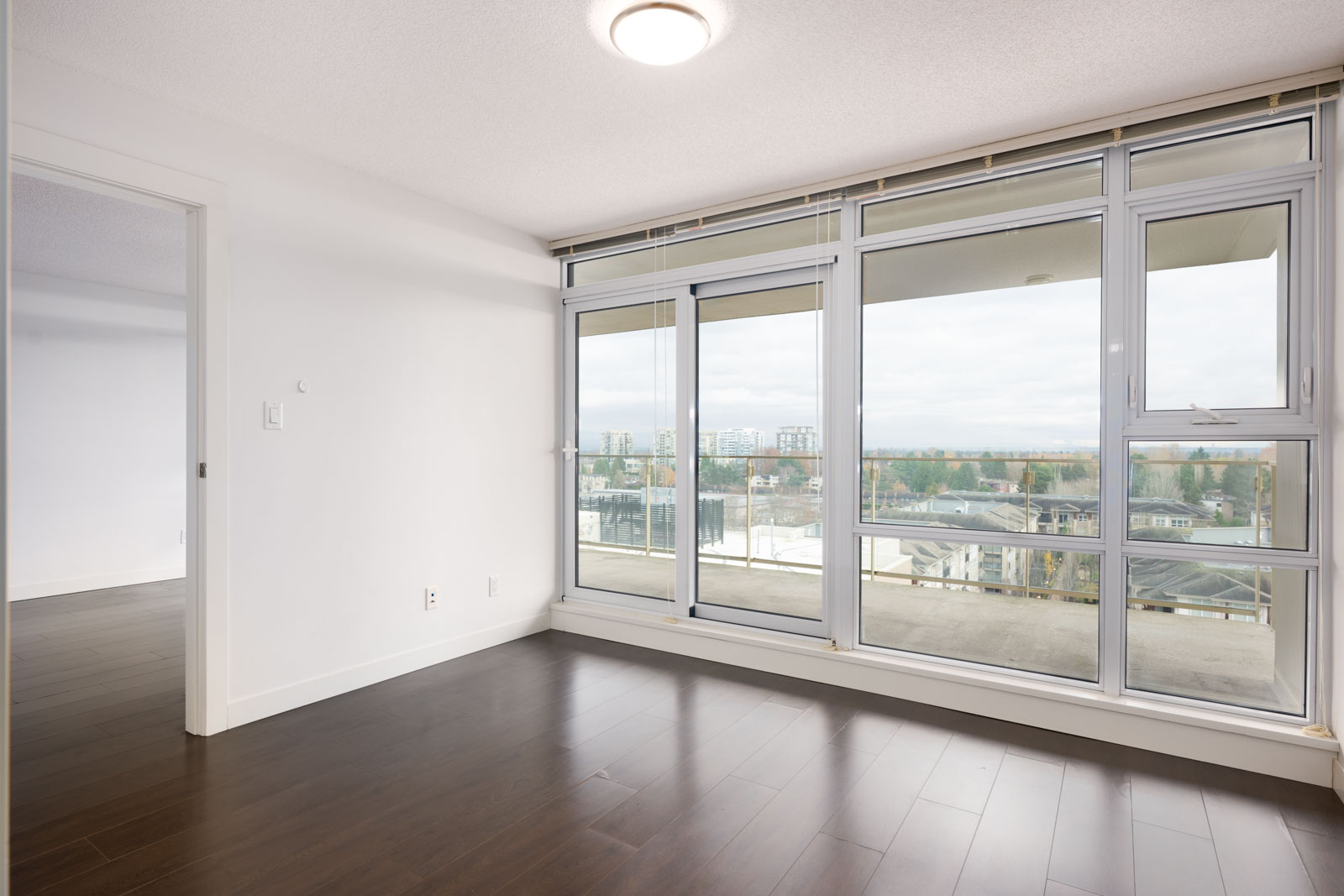 Unfurnished apartment room with large windows and a glass door leading to a balcony, featuring dark wood floors and white walls.