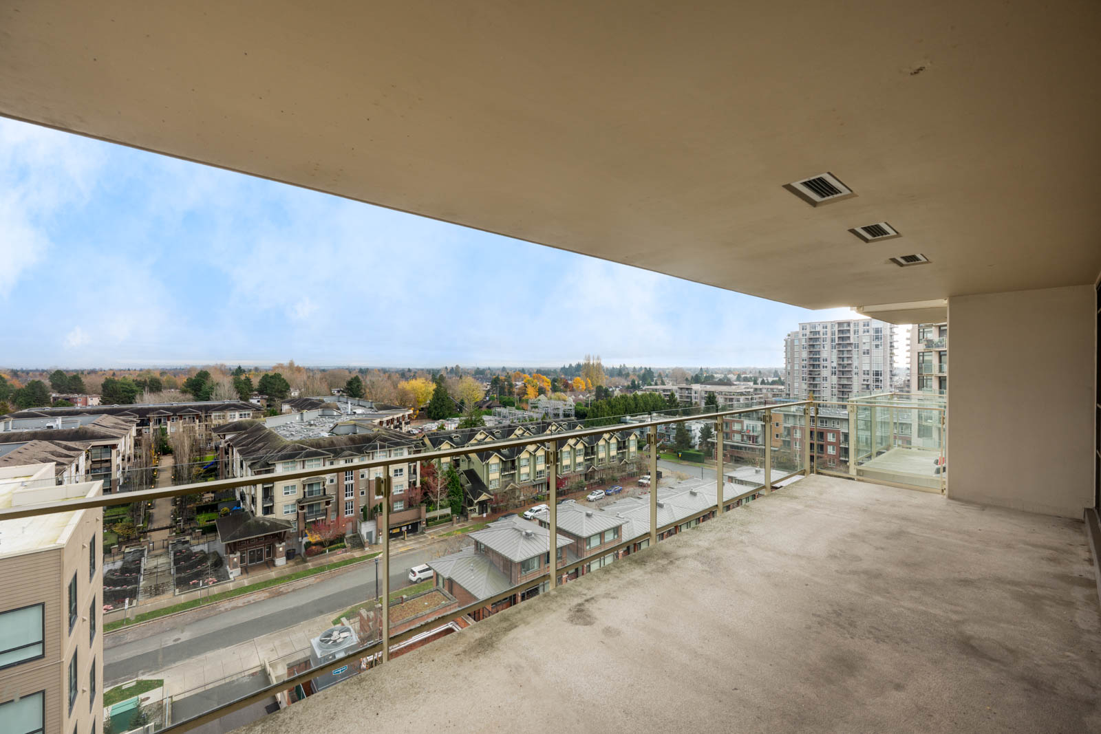 A spacious, empty balcony with glass railing overlooks a cityscape with mid-rise buildings, streets, and trees under a partly cloudy sky.
